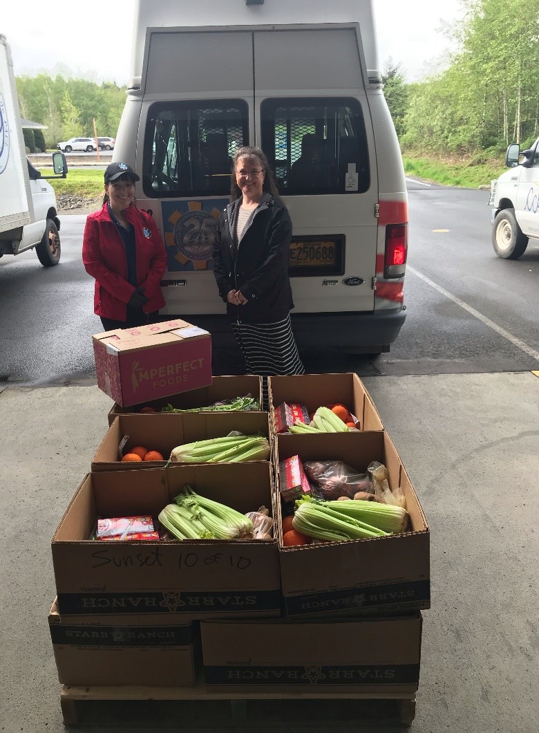 Image of food boxes containing celery, oranges and other fruits and veggies, stacked in front of a white van, delivered by the sunset team