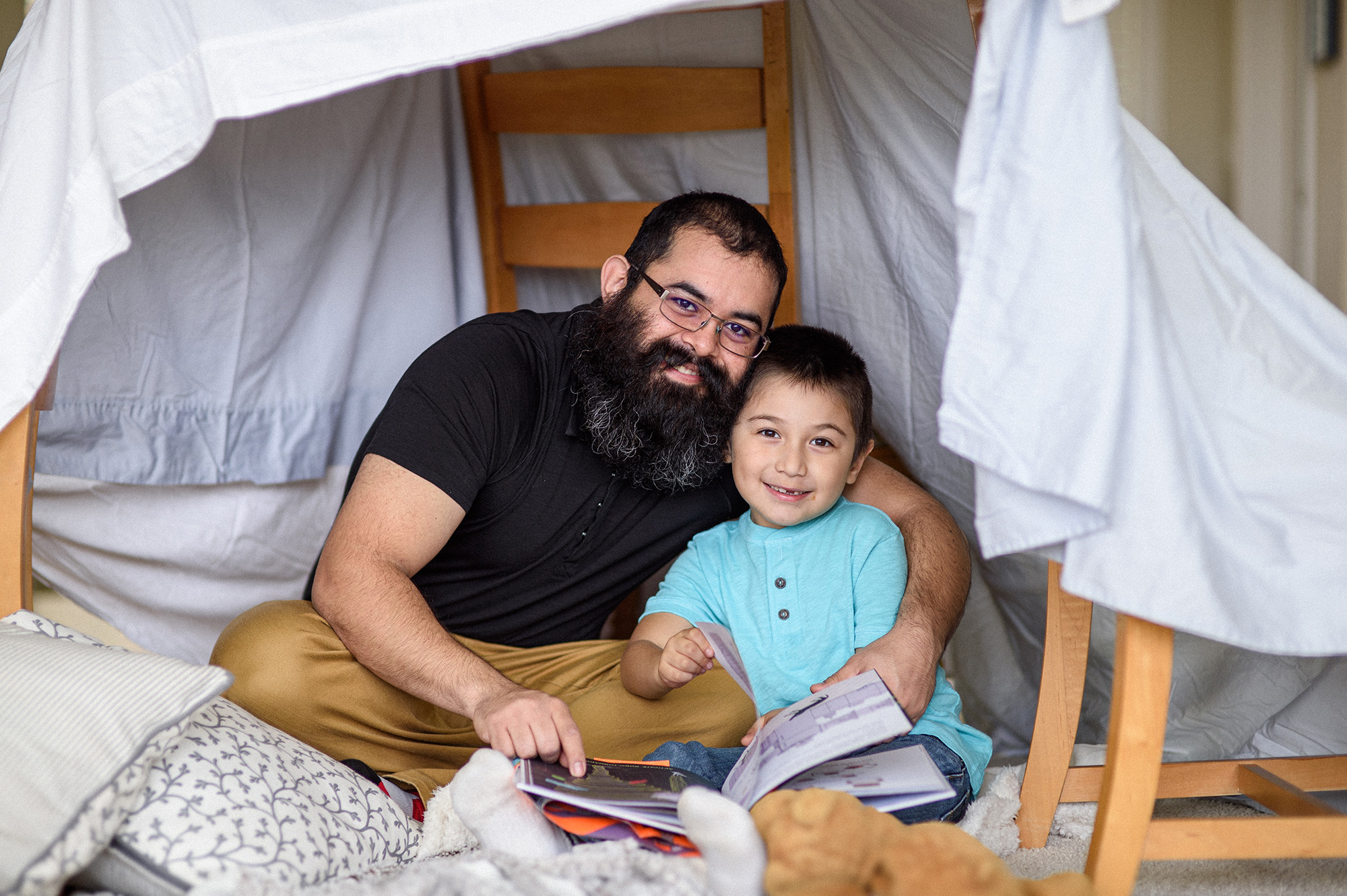 A father and son reading books under a fort made with a blanket and chairs